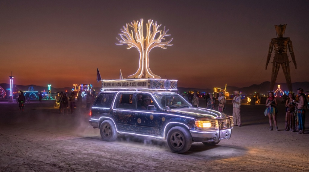 A colorful mutant vehicle decorated with lights and cosmic symbols drives across the dusty playa at Burning Man 2026 under the Axis Mundi theme